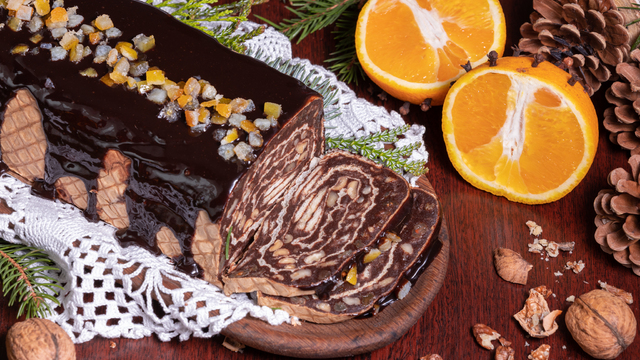 kakaowa rolada z wafli polana czekoladą, na niej kandyzowane owoce w kolorach pomarańczowym, niebieskim, żółtym. Rolada leży na desce do krojenia, ucięte są dwa kawałki, pod deką biała ludowa serwetka, obok leży gałązka świerku i dwie połówki pomarańczy