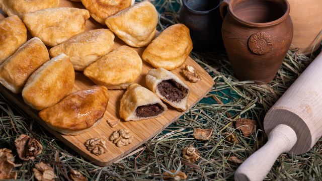 pieczone pierożki ułożone na desce do krojenia która leży na sianie i zielonym obrusie obok dwa puste gliniane garneczki