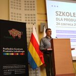 mężczyzna przy mówinicy w tle flagi polski i województwa i rollup z logiem Żubra i pcpl.jpg